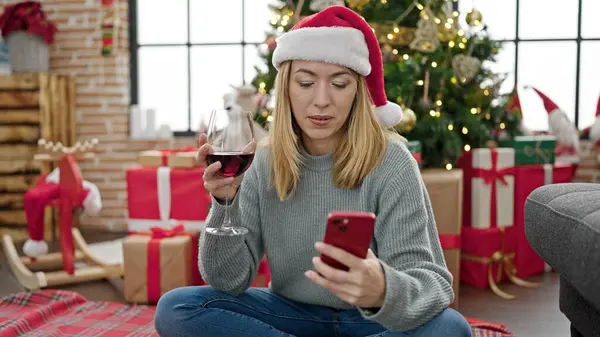 Young blonde woman using smartphone drinking wine celebrating christmas at home