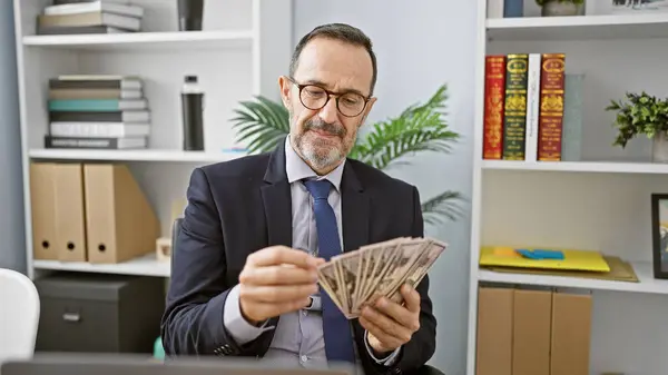 Beaming middle age man with grey hair, confident business worker, joyfully counting dollars at his office desk