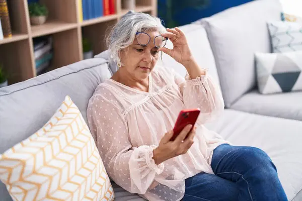 Middle age grey-haired woman looking screen smartphone with vision problem at home