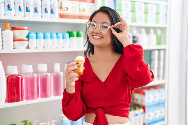 Young chinese woman customer smiling confident holding pills bottle at pharmacy