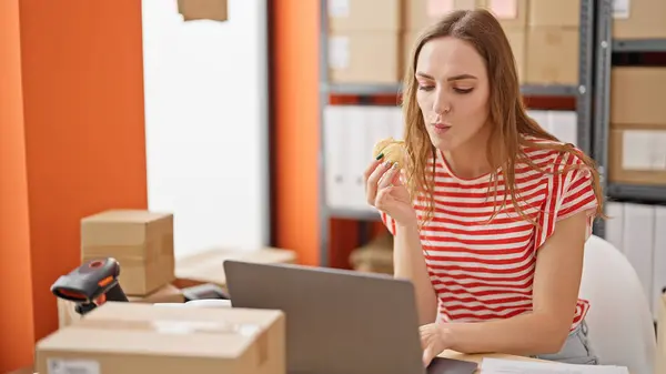 办公室使用笔记本电脑吃羊角面包的金发年轻女性电子商务工作者 — 图库照片