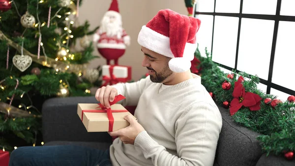Νεαρός Ισπανός Ξεπακετάρει Δώρο Καθισμένος Στον Καναπέ Δίπλα Στο Χριστουγεννιάτικο — Φωτογραφία Αρχείου