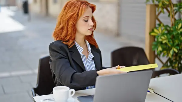 Young redhead woman business worker holding documents of binder at coffee shop terrace