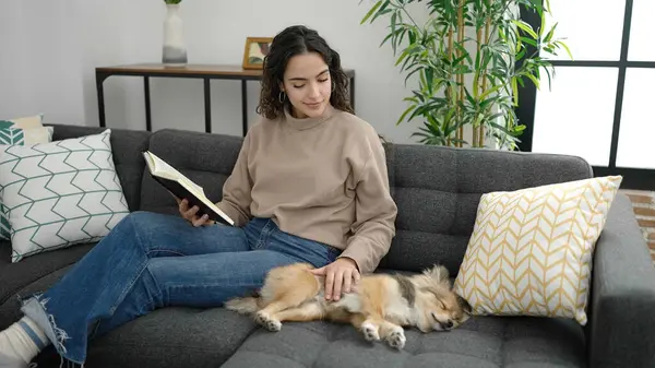 Young hispanic woman with dog reading book sitting on sofa at home