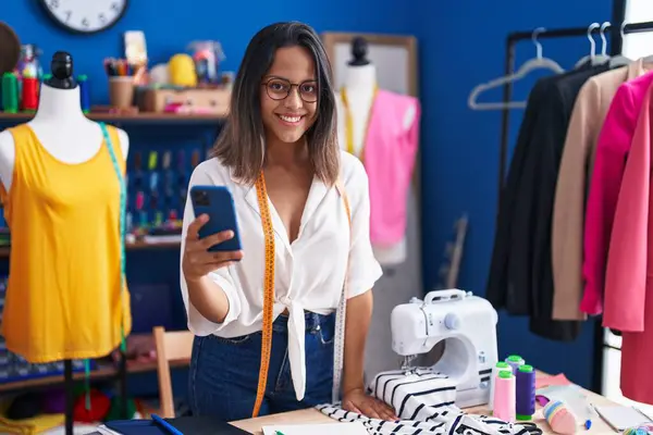 Young hispanic woman tailor smiling confident using smartphone at sewing studio