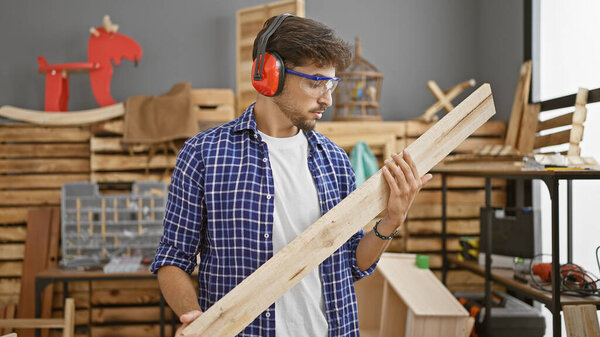 Handsome young arab man absorbed in his profession, skillfully handling his wood plank at a cozy indoor carpentry workshop.