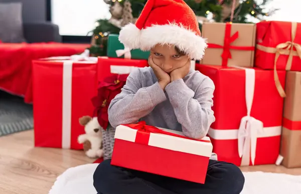自宅で不幸な表現で床に座っているクリスマスの贈り物を保持する好ましいヒスパニックの少年 — ストック写真
