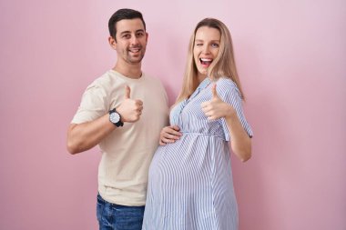 Pembe arka planda bebek bekleyen genç bir çift elleriyle mutlu bir jest yapıyor. Kameraya bakıp başarıyı gösteren ifade. 