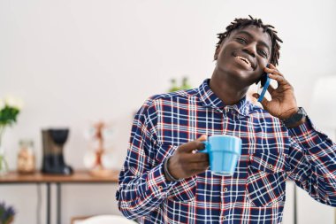 Afro-Amerikalı adam akıllı telefondan konuşuyor evde kahve içiyor.