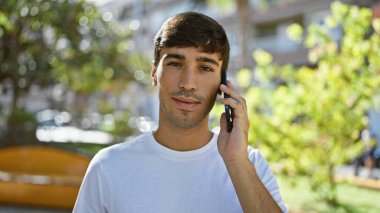 Serin, akıllı telefondan ciddi bir konuşma yapan İspanyol genç adam güneşli açık havanın altındaki yeşil şehir parkında rahatlamış ama ciddi bir yaşam tarzını ifade ediyor..