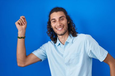 Young hispanic man standing over blue background dancing happy and cheerful, smiling moving casual and confident listening to music 