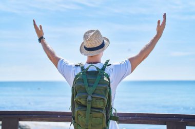 Young caucasian man tourist standing with arms open on back view at seaside