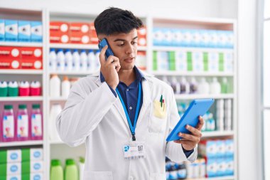 Young hispanic man pharmacist using touchpad talking on smartphone at pharmacy