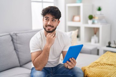 Sakallı İspanyol bir adam kanepeye oturmuş çene üstüne oturmuş soru soruyor, düşünceli bir yüz ifadesi var. Gülümseyen ve düşünceli bir yüz. şüphe kavramı. 