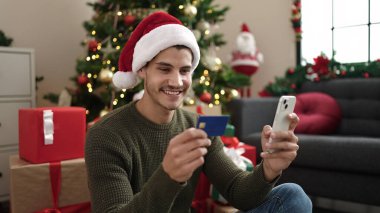 Genç İspanyol adam akıllı telefon ve kredi kartı kullanıyor. Evdeki Noel ağacının yanında yerde oturuyor.