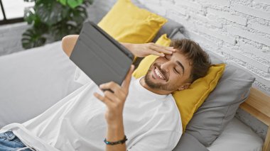 Yatak odasının rahatlığında, genç, gülümseyen bir Arap adam yatağa uzanıyor, dokunmatik bilgisayarında bir video seyrediyor, her anın tadını çıkarıyor..