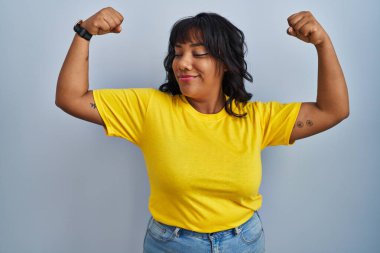 Mavi arka planda duran İspanyol kadın gururla gülümseyen kol kaslarını gösteriyor. fitness konsepti. 