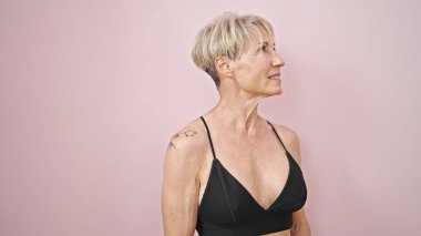 Orta yaşlı, kendine güvenen, pembe arka planda bikini giyen sarışın bir kadın.