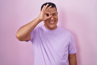 Young hispanic man standing over pink background doing ok gesture with hand smiling, eye looking through fingers with happy face. 