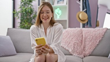 Kanepede oturmuş akıllı telefon kullanan genç sarışın kadın evde gülümsüyor.