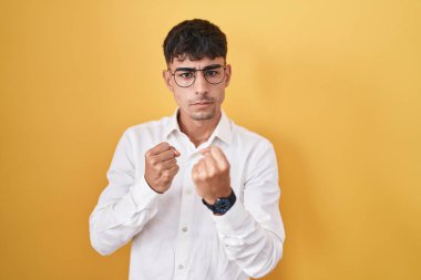 Young hispanic man standing over yellow background ready to fight with fist defense gesture, angry and upset face, afraid of problem 