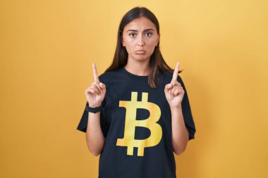 Bitcoin tişörtü giyen genç İspanyol kadın üzgün ve üzgün görünüyor, parmaklarıyla yönü gösteriyor, mutsuz ve depresif.. 