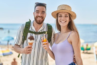 Kadın ve erkek turist, deniz kenarında dondurma yiyerek gülümsüyor.