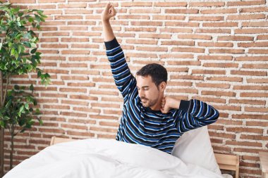 Young hispanic man waking up stretching arms at bedroom