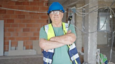 Orta yaşlı, gri saçlı, gülümseyen, kendine güvenen, inşaat alanında kollarını kavuşturup el hareketi yapan bir adam.