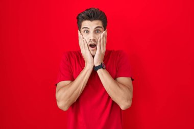 Young hispanic man standing over red background afraid and shocked, surprise and amazed expression with hands on face 