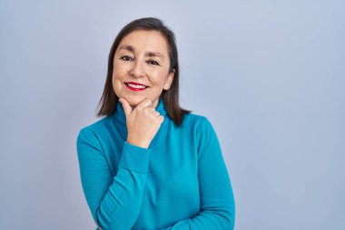 Middle age hispanic woman standing over isolated background looking confident at the camera smiling with crossed arms and hand raised on chin. thinking positive. 