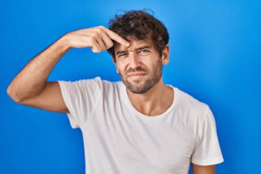 Mavi arka planda duran İspanyol genç adam alnındaki sivilceden mutsuz olduğunu gösteriyor, siyah başlıklı çirkin bir enfeksiyon. sivilce ve deri problemi 