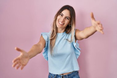 Pembe arka planda duran genç İspanyol kadın kameraya sarılmak için kollarını açıp gülümseyerek bakıyor. Mutluluğu kucaklayan neşeli bir ifade.. 