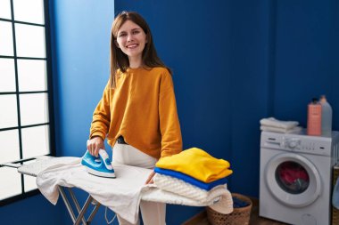 Genç sarışın kadın gülümsüyor çamaşırhanede kendinden emin ütü yapıyor.