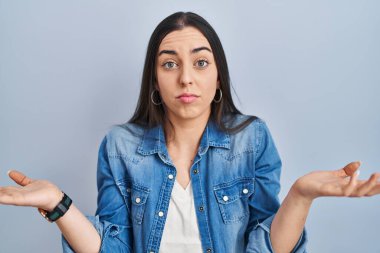 Hispanic woman standing over blue background clueless and confused expression with arms and hands raised. doubt concept. 