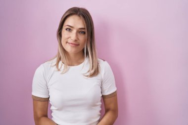 Blonde caucasian woman standing over pink background with hands together and crossed fingers smiling relaxed and cheerful. success and optimistic 