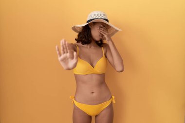 Genç İspanyol kadın bikini giyiyor ve yaz şapkası takıyor. Gözleri ellerle örtüyor ve üzgün ve korku ifadeleriyle jest yapıyor. utangaç ve olumsuz kavram. 