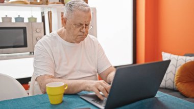 Middle age grey-haired man using laptop with relaxed expression at dinning room
