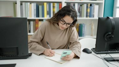 Genç ve güzel İspanyol kız öğrenci kütüphanede not tutarak akıllı telefon kullanıyor.