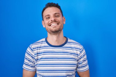 Mavi arka planda duran genç İspanyol adam ellerini birleştirip gülümseyerek gevşemiş ve neşeliydi. Başarı ve iyimserlik 