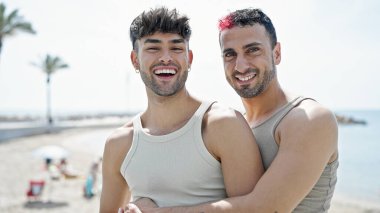 İki erkek, deniz kenarında birbirlerine sarılarak gülümsüyor.