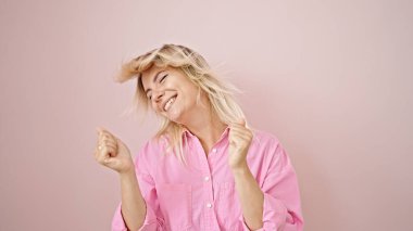 Genç sarışın kadın gülümsüyor pembe arka planda dans ediyor.