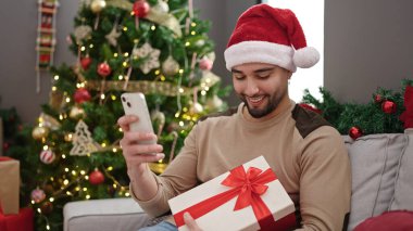 Genç Arap adam elinde akıllı telefon hediyesiyle evde Noel ağacının yanında oturuyor.