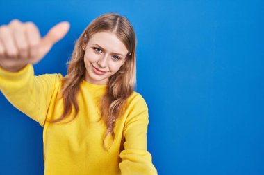 Mavi arka planda durup olumlu bir jest yapmayı onaylayan, gülümseyen ve başarı için mutlu genç beyaz bir kadın. kazanan jest. 