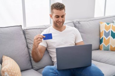 Evdeki kanepede oturan genç, beyaz bir adam dizüstü bilgisayar ve kredi kartı kullanıyor.