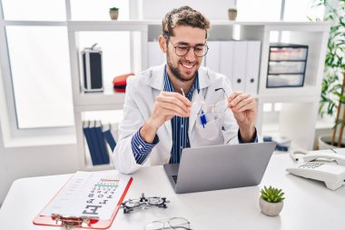 Young man optician holding glasses using laptop at clinic