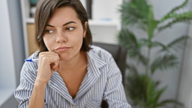 İş yerinde, genç, güzel İspanyol kadın çalışan, ciddi düşüncelerle büyülenmiş, ofis masasında oturan, iş başarısına odaklanmış bir portre..