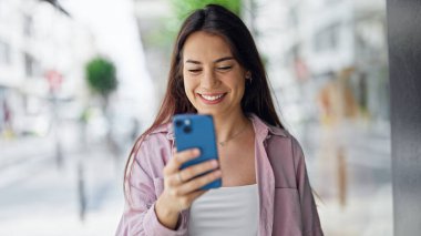 Genç, güzel, İspanyol bir kadın sokakta akıllı telefon kullanarak gülümsüyor.