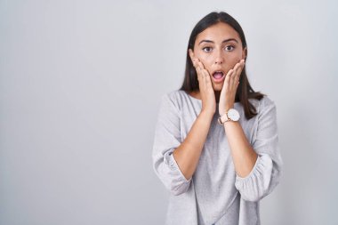 Young hispanic woman standing over white background afraid and shocked, surprise and amazed expression with hands on face 