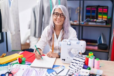 Orta yaşlı, gri saçlı, sırıtan, kendine güvenen, terzi dükkanındaki defterine çizim yapan bir kadın.